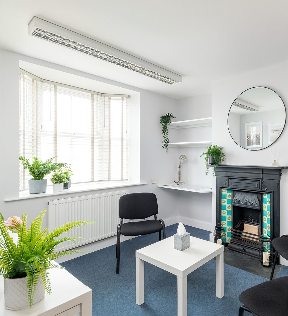 The calm in-person room at the Barnstaple Chiropractic & Complementary Health Centre in Sticklepath — a bright bay window with blinds, plants, a comfortable chair and a traditional fireplace