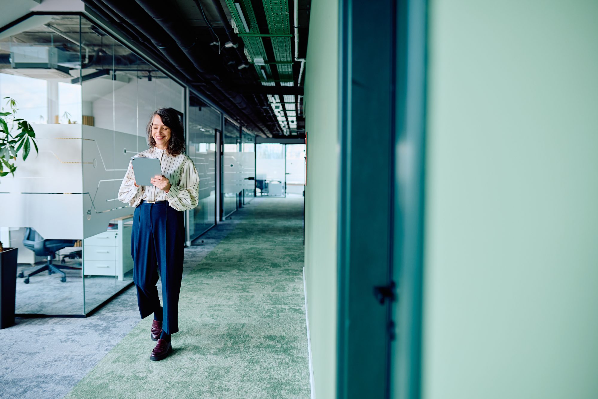 A smiling professional walking through a bright, green-toned modern office while reading from a tablet